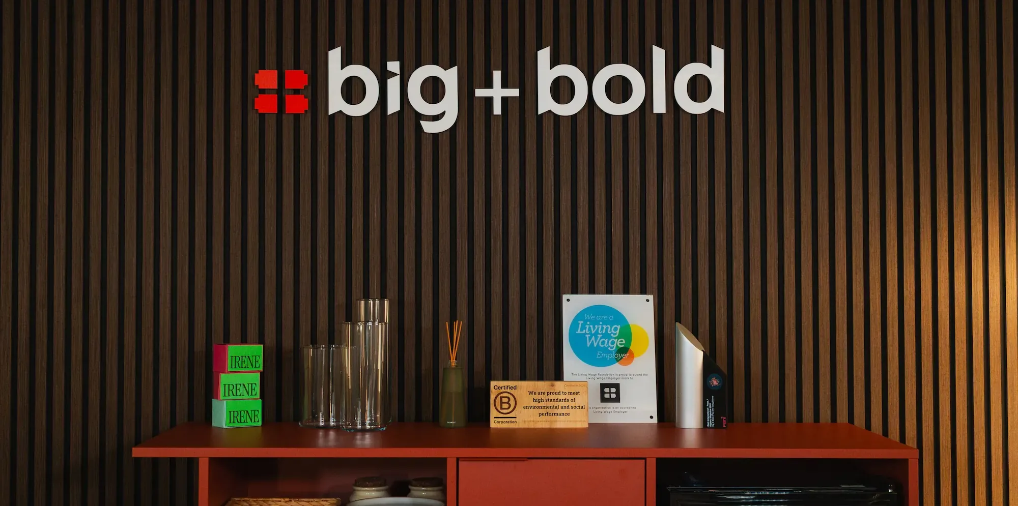 The big+bold logo on a slatted wooden wall in the studio, above a red sideboard with the IRENE blocks and the B Corp and Living Wage plaques.