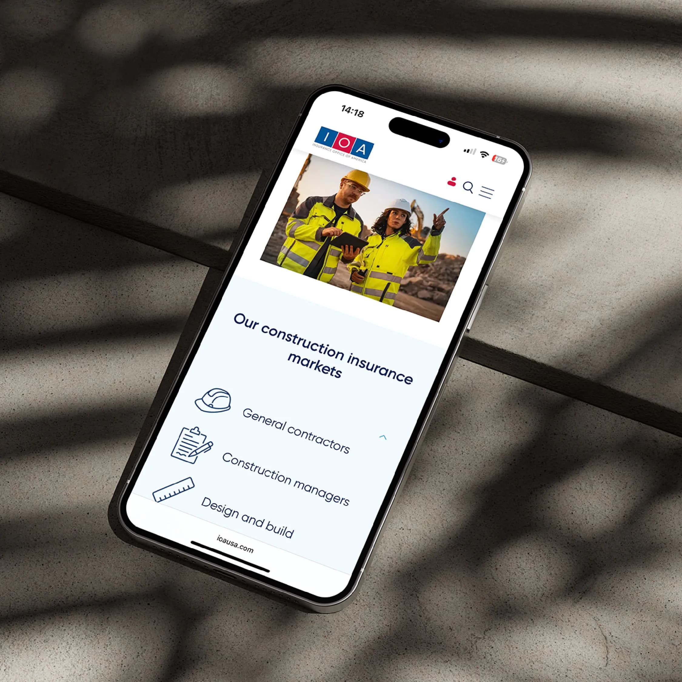 A phone in dappled shadow shows IOA’s construction insurance page, listing general contractors, construction managers, and design and build.