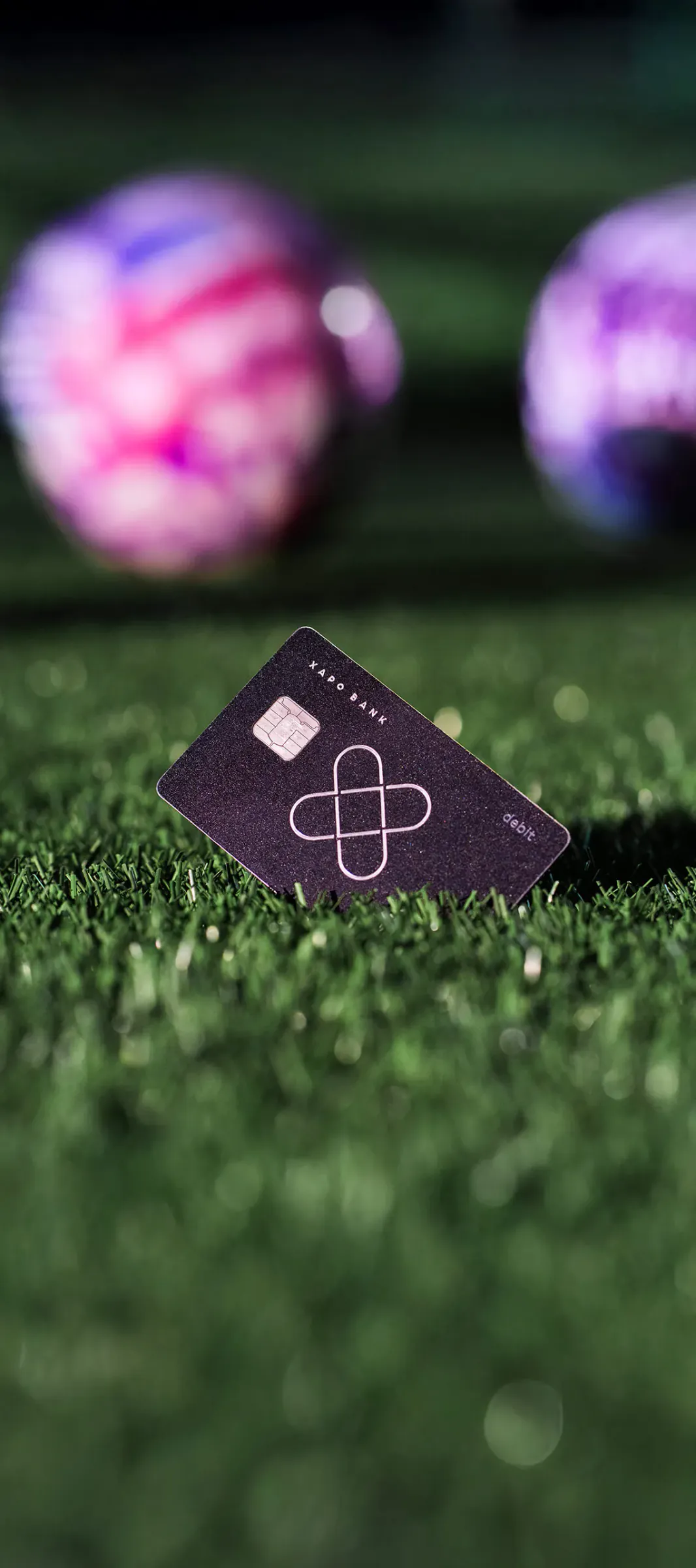 A black Xapo Bank debit card stands on floodlit turf, with two match balls glowing pink and purple behind it.