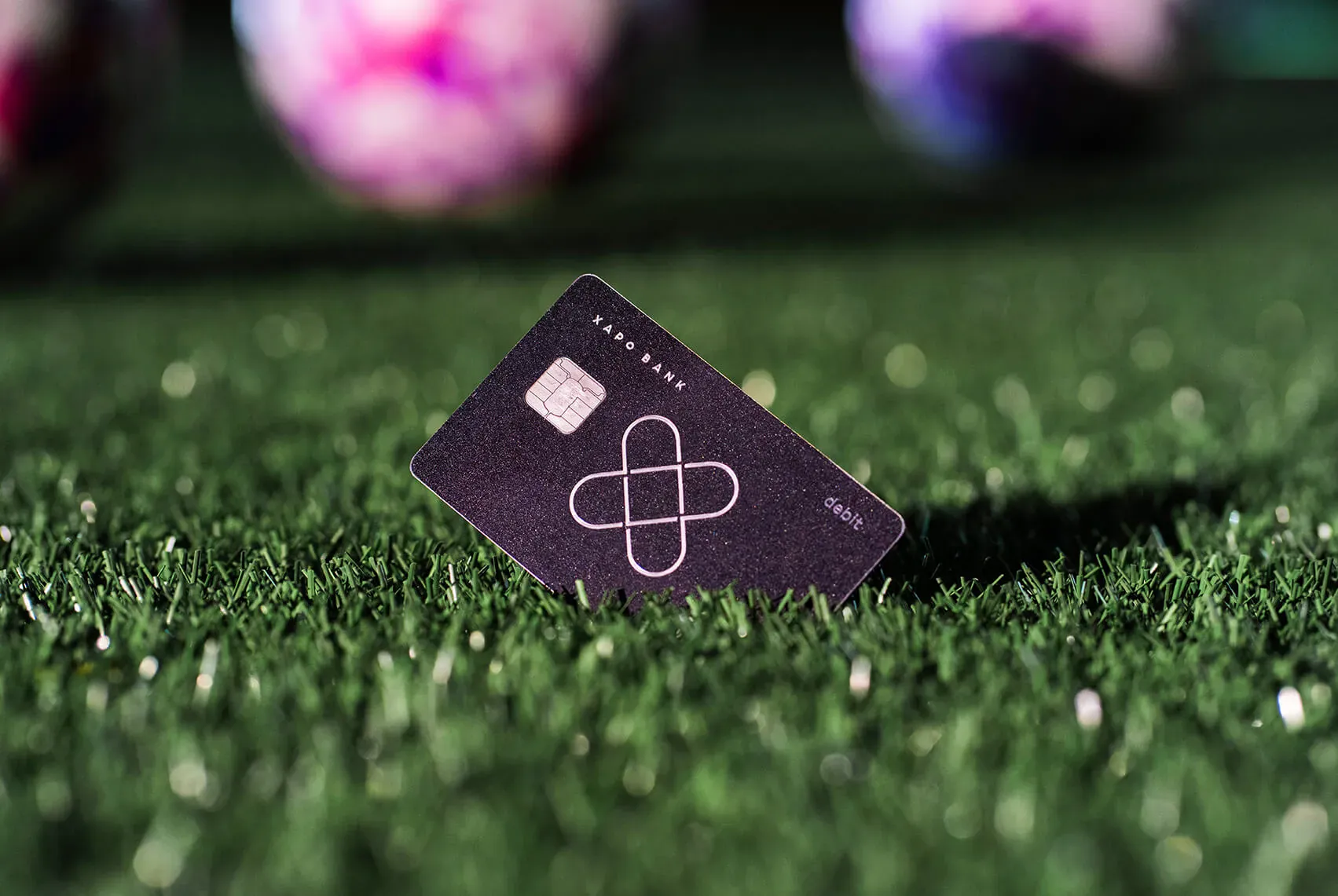 A black Xapo Bank debit card stands on floodlit turf, with two match balls glowing pink and purple behind it.