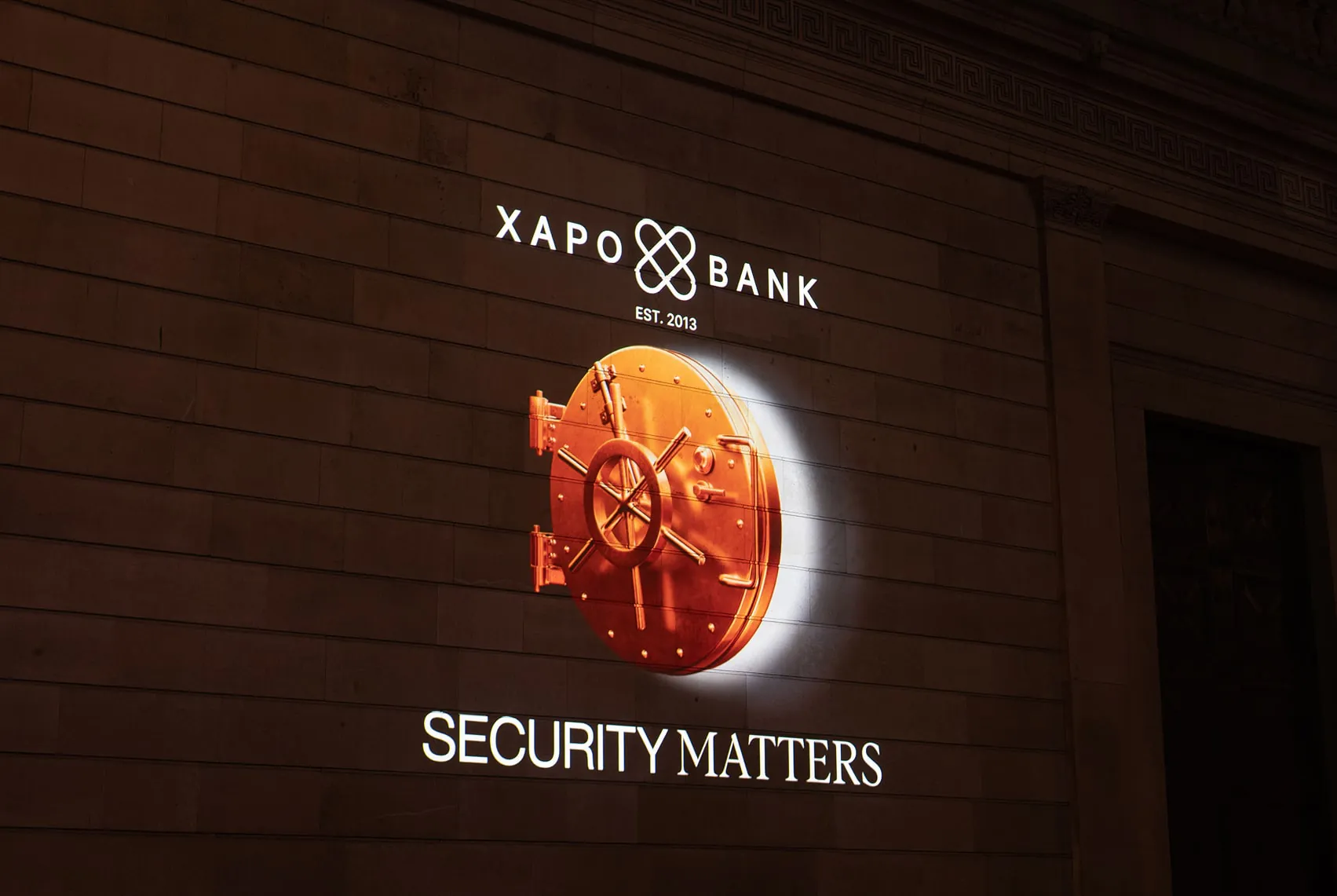 The Xapo Bank logo, a glowing copper vault door and “Security Matters” projected onto a brick wall at night.